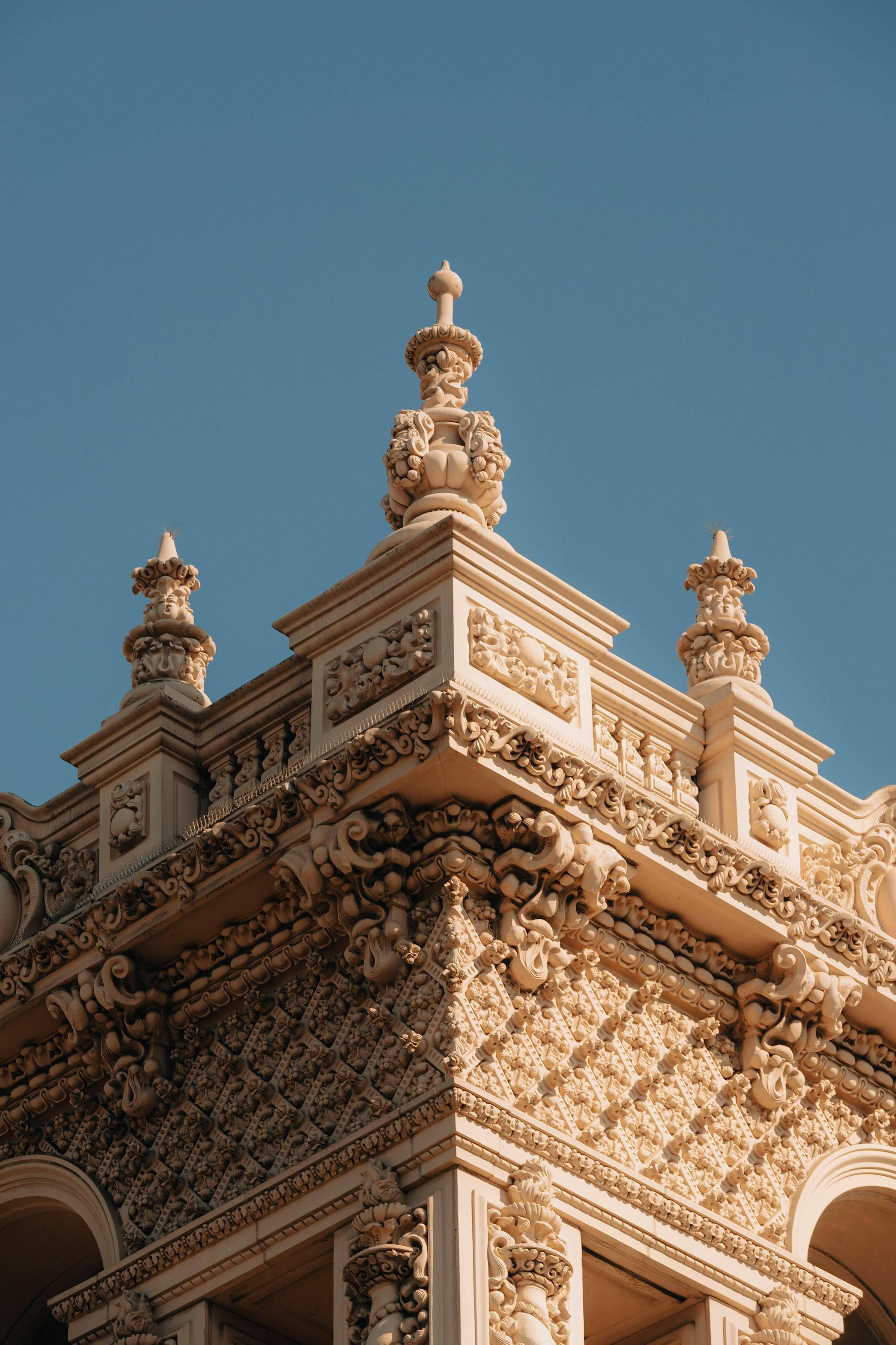 Close-up of ornate cream-colored architectural corner with detailed carvings and finials against blue sky.