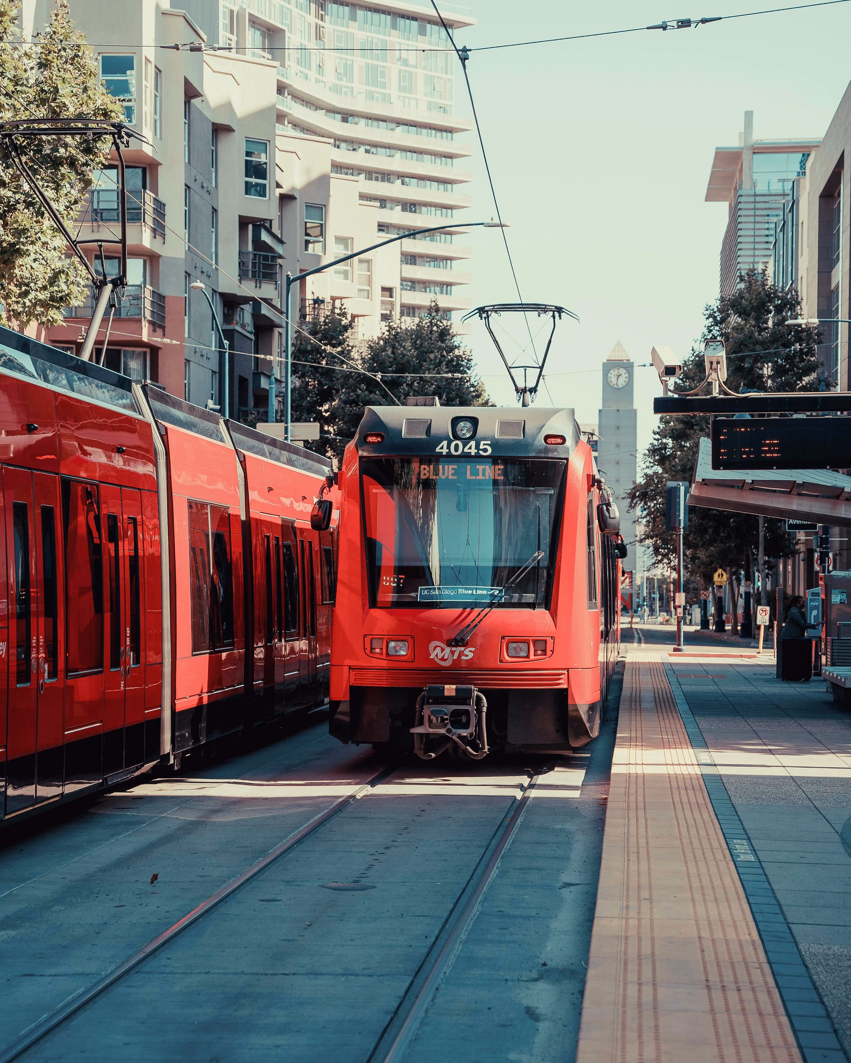 Red MTS Blue Line light rail train arriving at an urban station with tracks and platform visible.