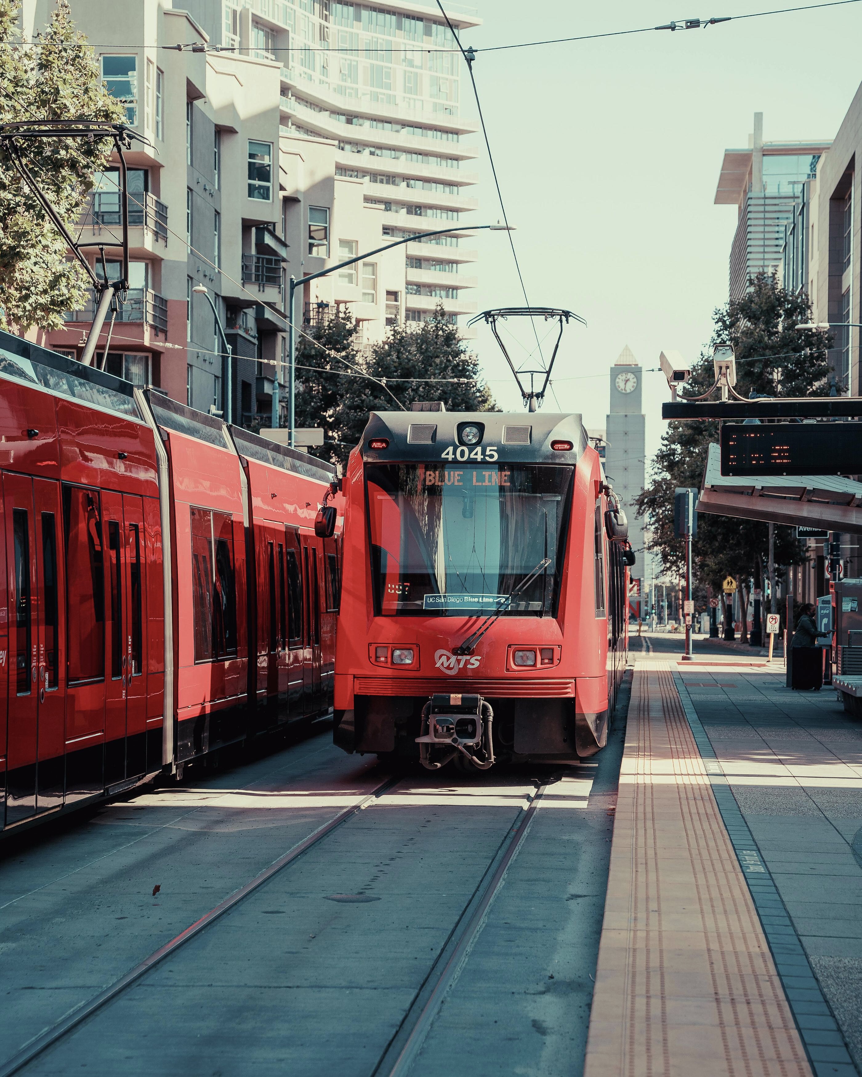 Red MTS Blue Line light rail train arriving at an urban station with tracks and platform visible.