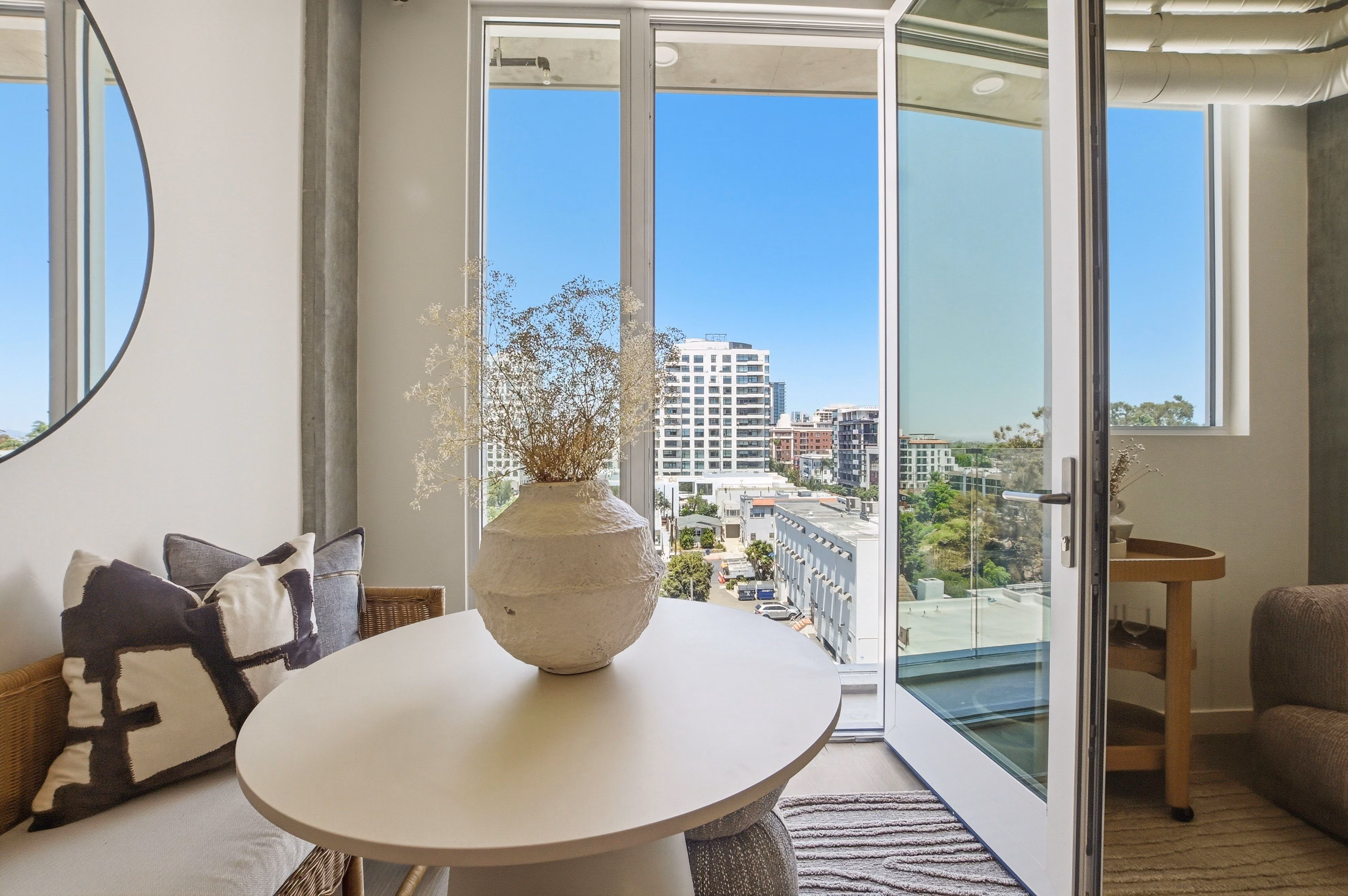 Modern room with round table, vase, cushions, and open glass door overlooking city buildings.
