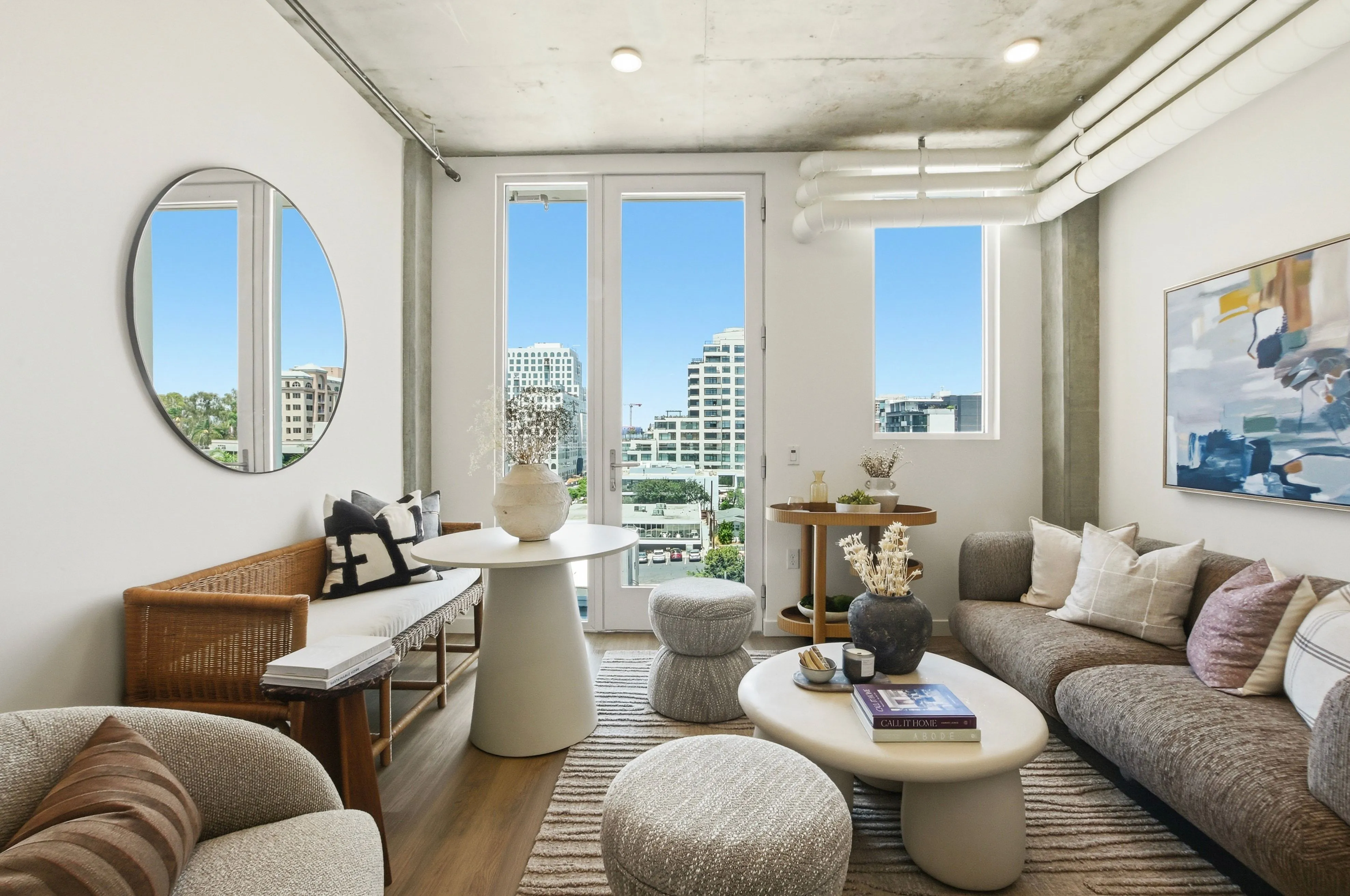 Modern living room with sofa, wicker bench, coffee tables, poufs, abstract art, and city view windows.