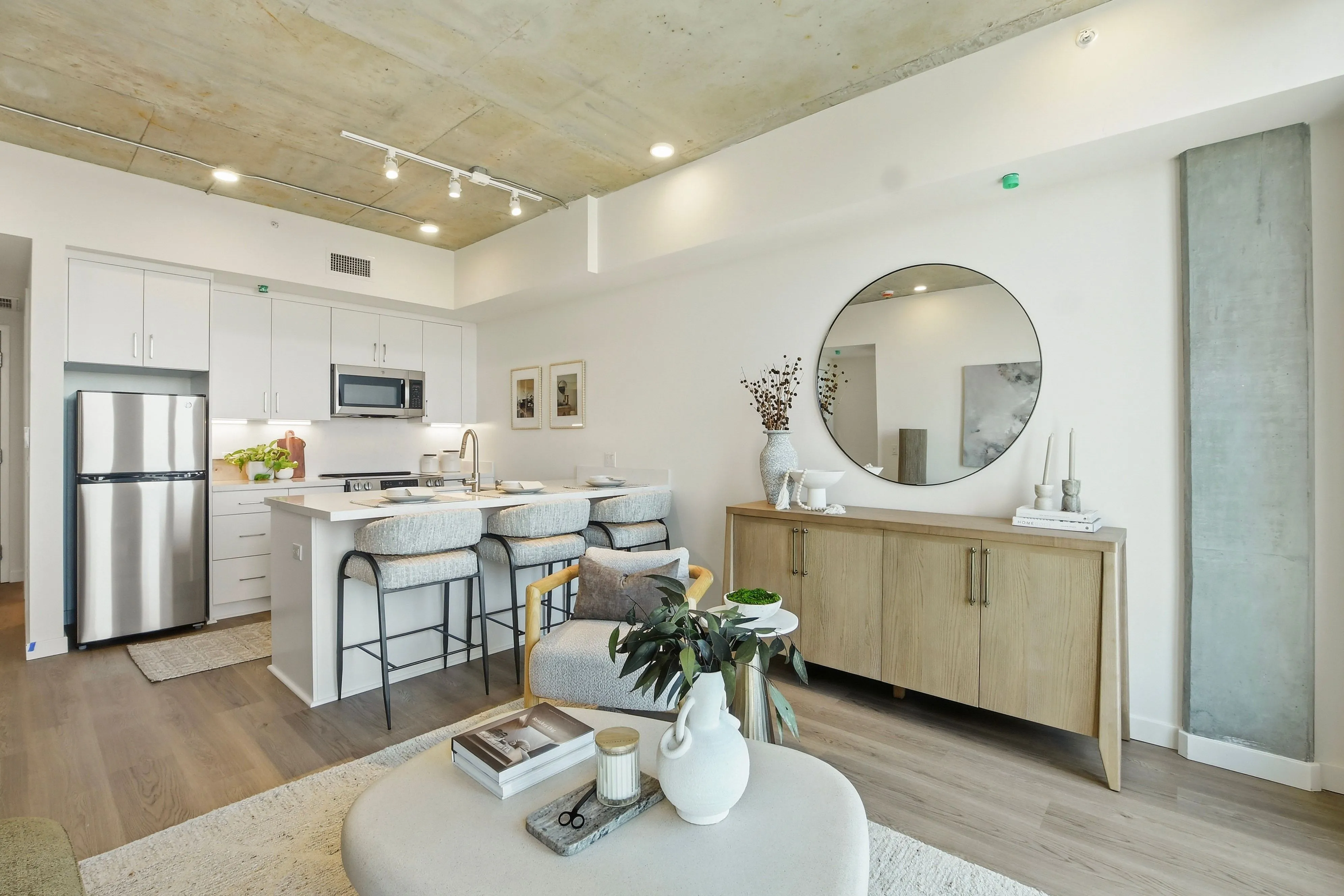 Modern open kitchen and living area with white cabinets, bar stools, wooden sideboard, and round mirror.