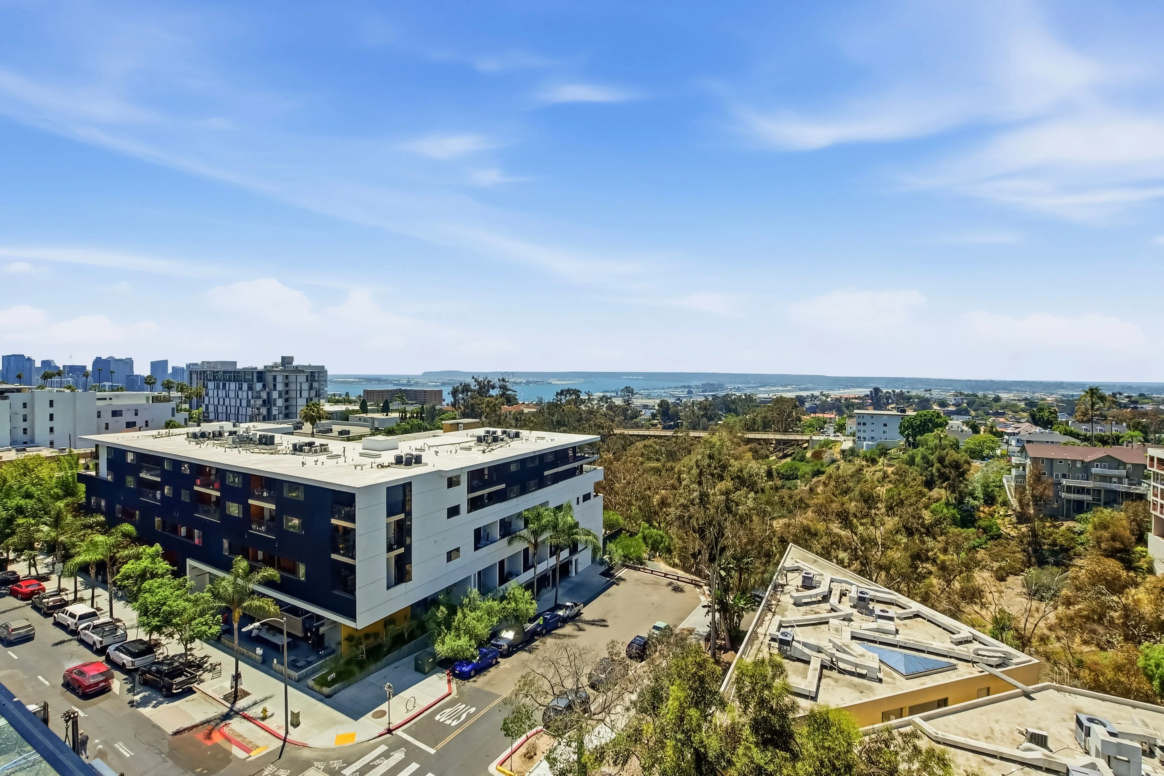 Urban area with modern apartment buildings, lush green trees, and a distant view of the ocean under blue sky.