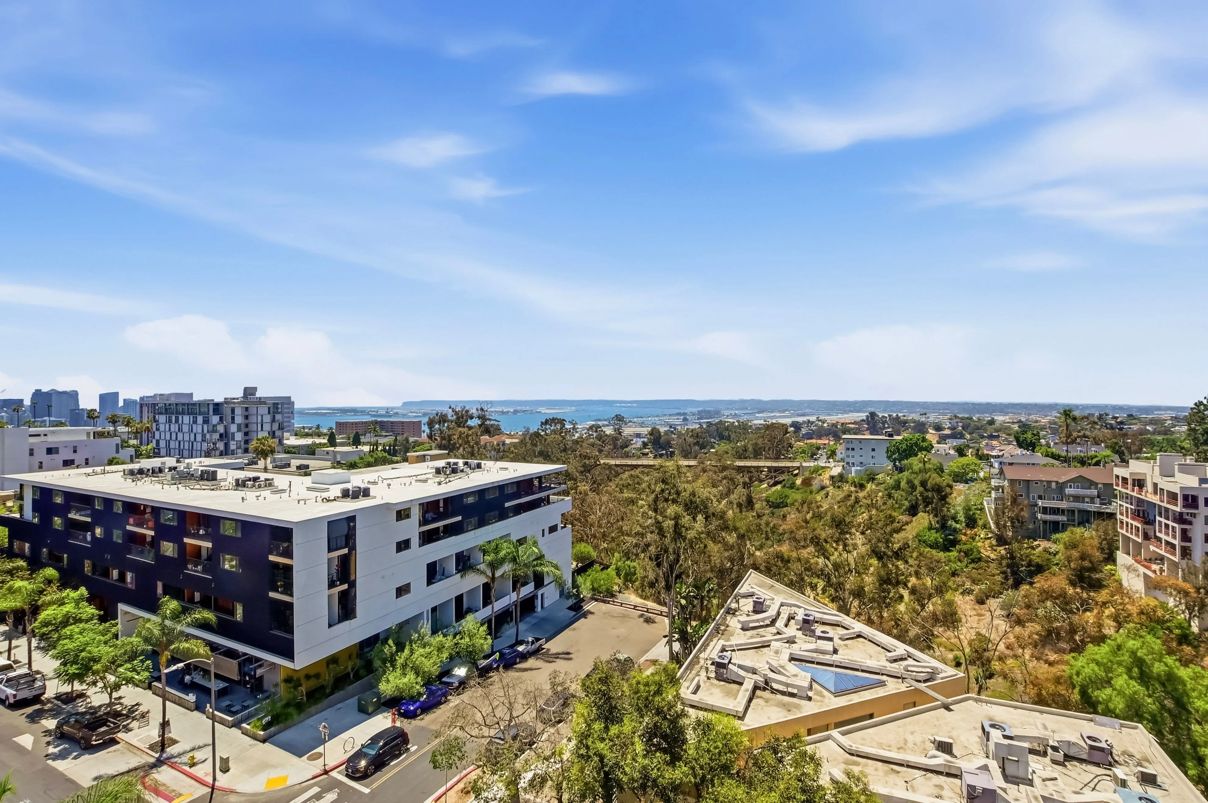 Urban neighborhood with apartment buildings, trees, streets, and distant water under a blue sky.