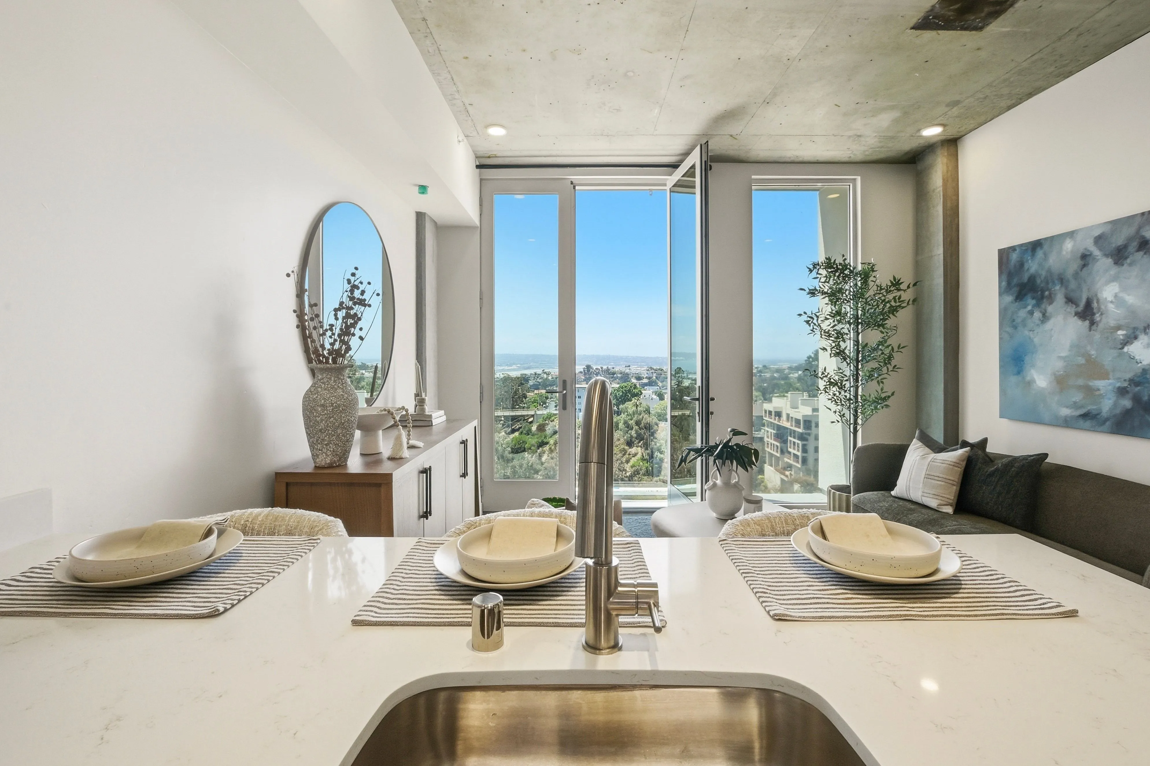 Modern kitchen island with sink and place settings, opening to a living room with glass doors and city view.