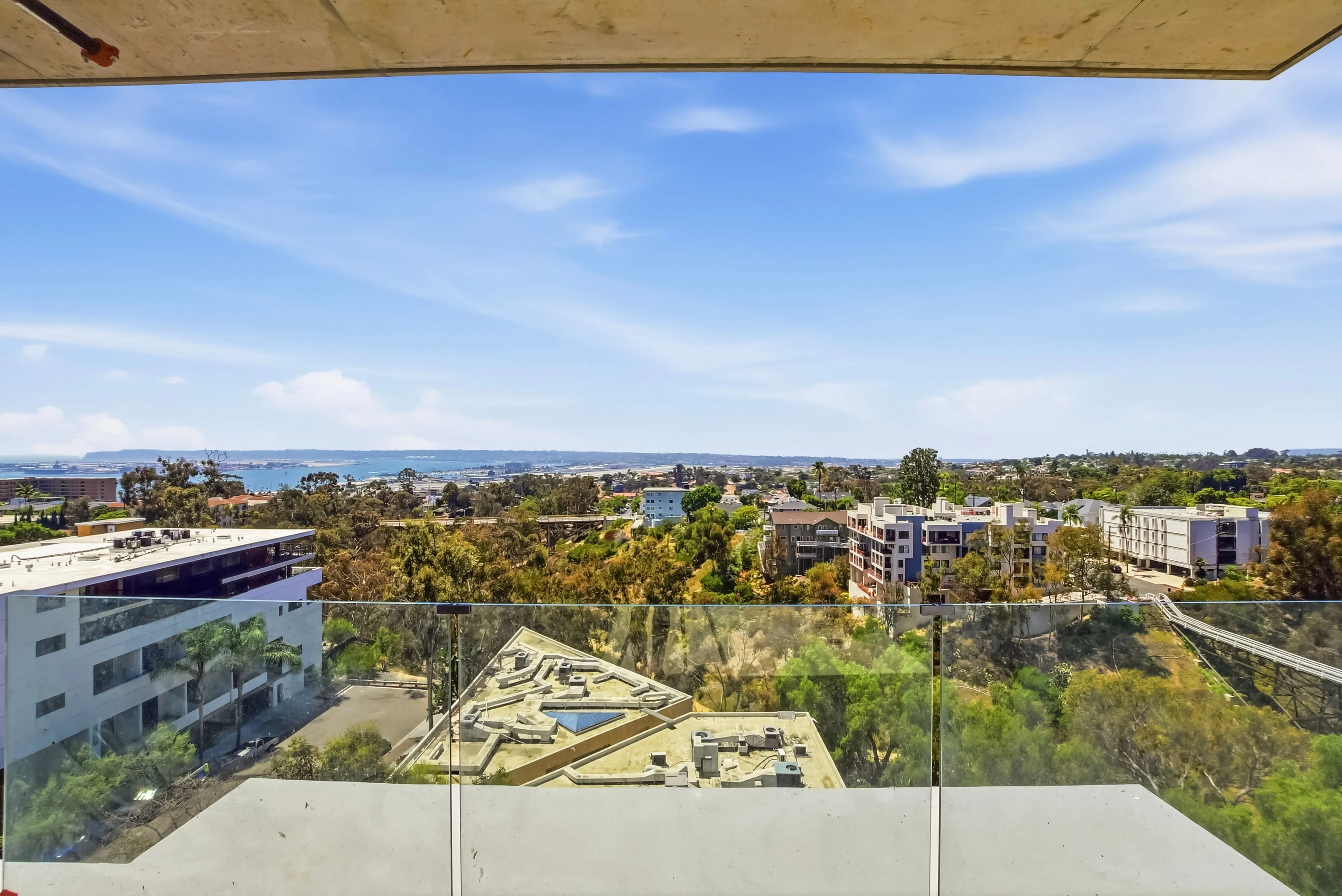 View from a balcony with glass railing overlooking buildings, trees, and a distant body of water.