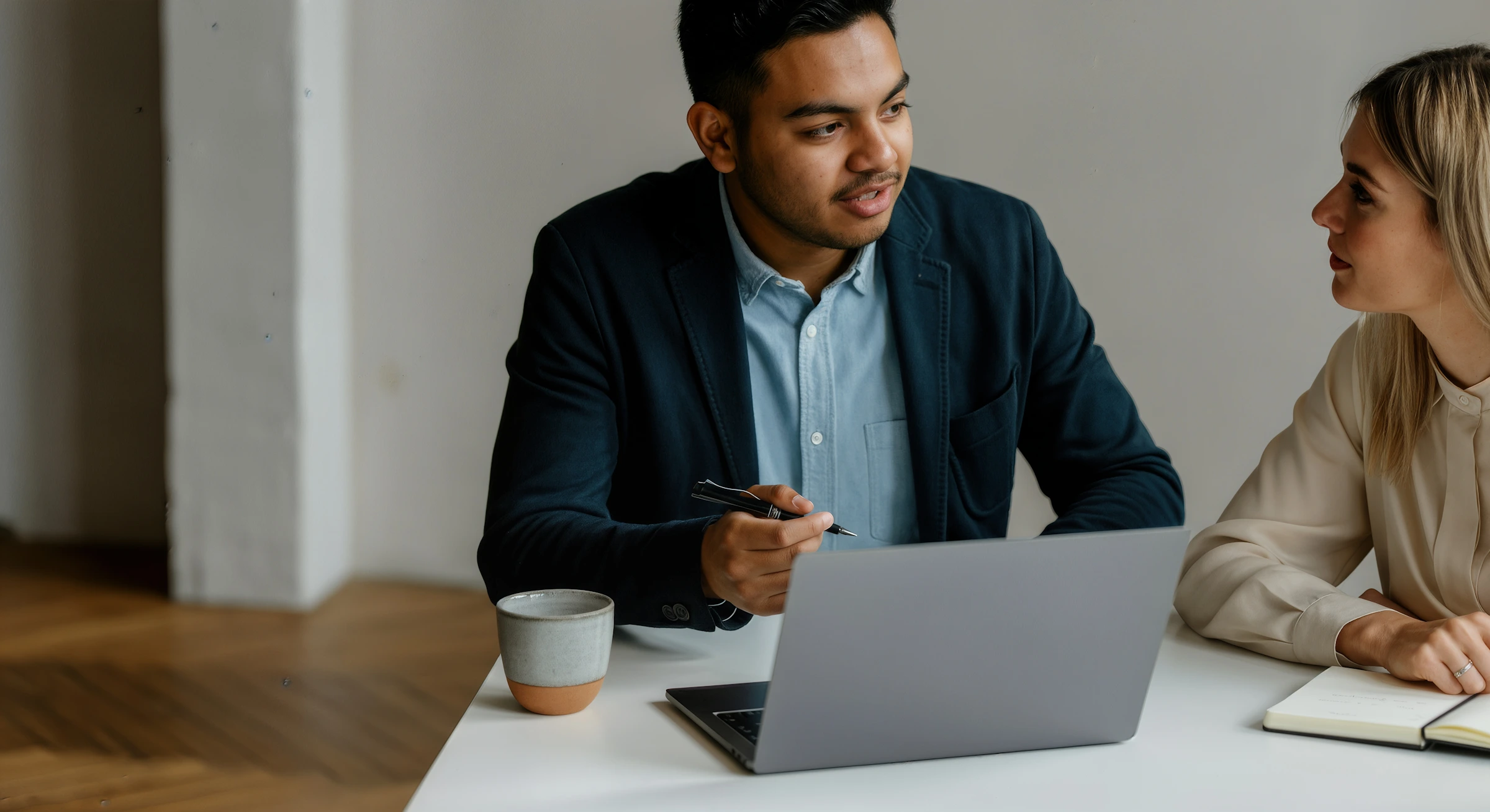 Man in a dark blazer holding a pen talking to a woman in a beige blouse at a desk with a laptop and notebook.