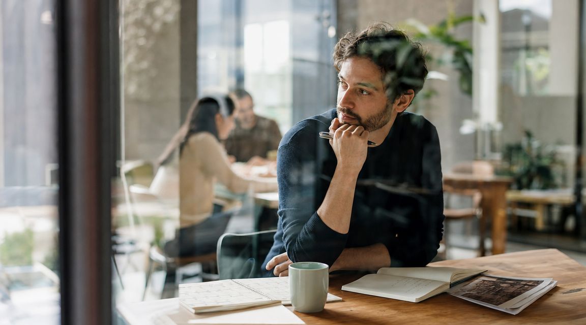 A pensive young man sitting at a desk with a notebook and coffee, looking away thoughtfully.