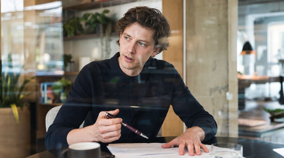 A professional man sitting at a desk with a pen, looking off-camera during a business meeting.