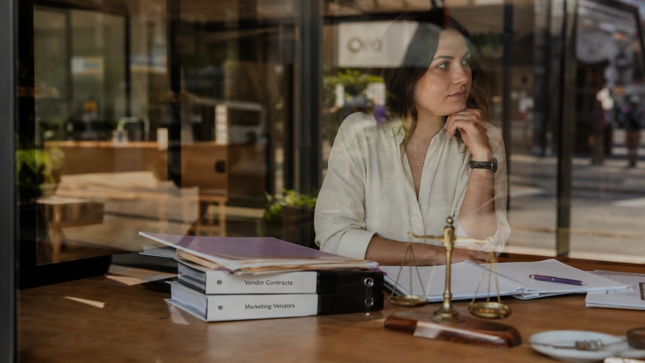 A professional woman sitting at a desk with vendor contract binders and a decorative scale of justice.