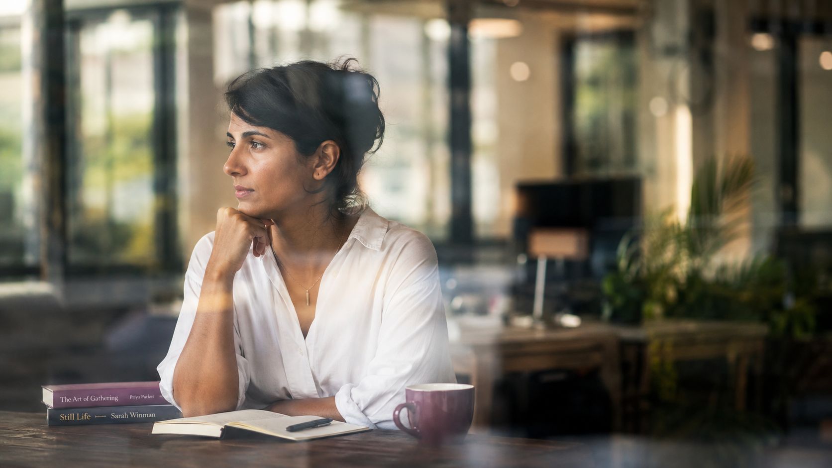 A contemplative woman sitting at a table with books and a coffee mug in an office setting.
