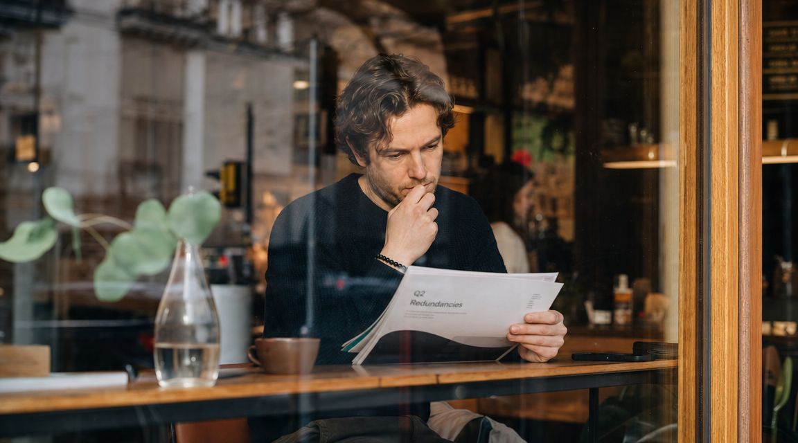 A focused man sitting at a cafe table looking at a report titled Q2 Redundancies.