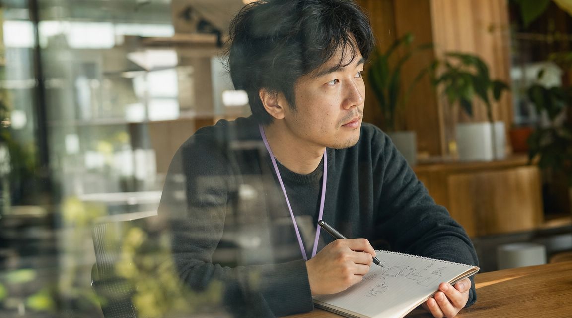 A focused Asian man wearing a lanyard sits at a desk drawing a diagram in his notebook.