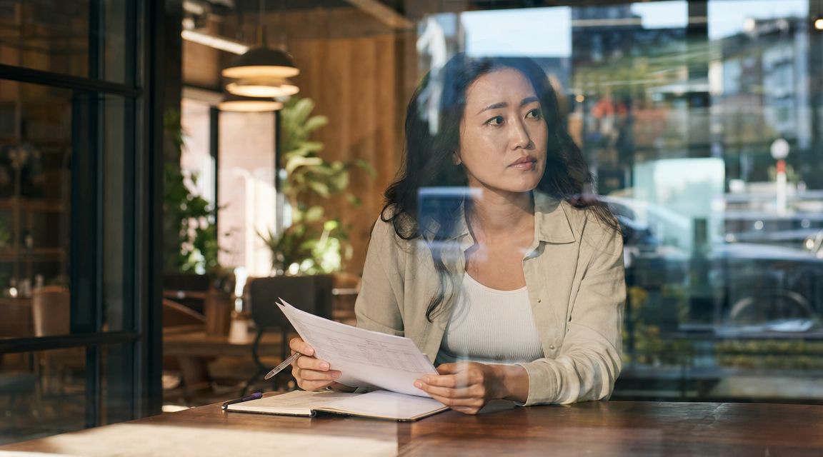 A contemplative woman sitting by a window reviewing documents as part of the procurement life cycle process.