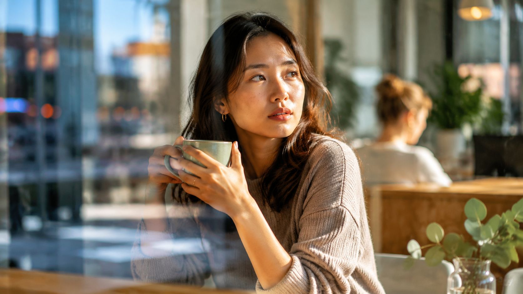 A young woman sits thoughtfully in a cafe by a window, holding a warm mug of coffee.