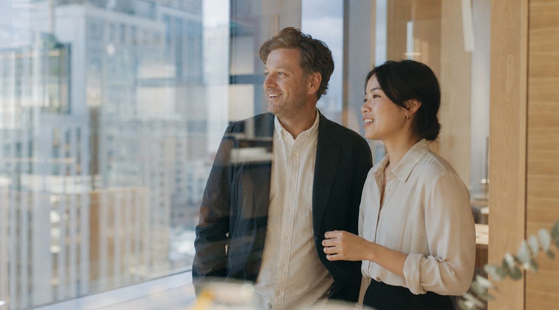 A professional man and woman standing together in an office, looking out of a large glass window.