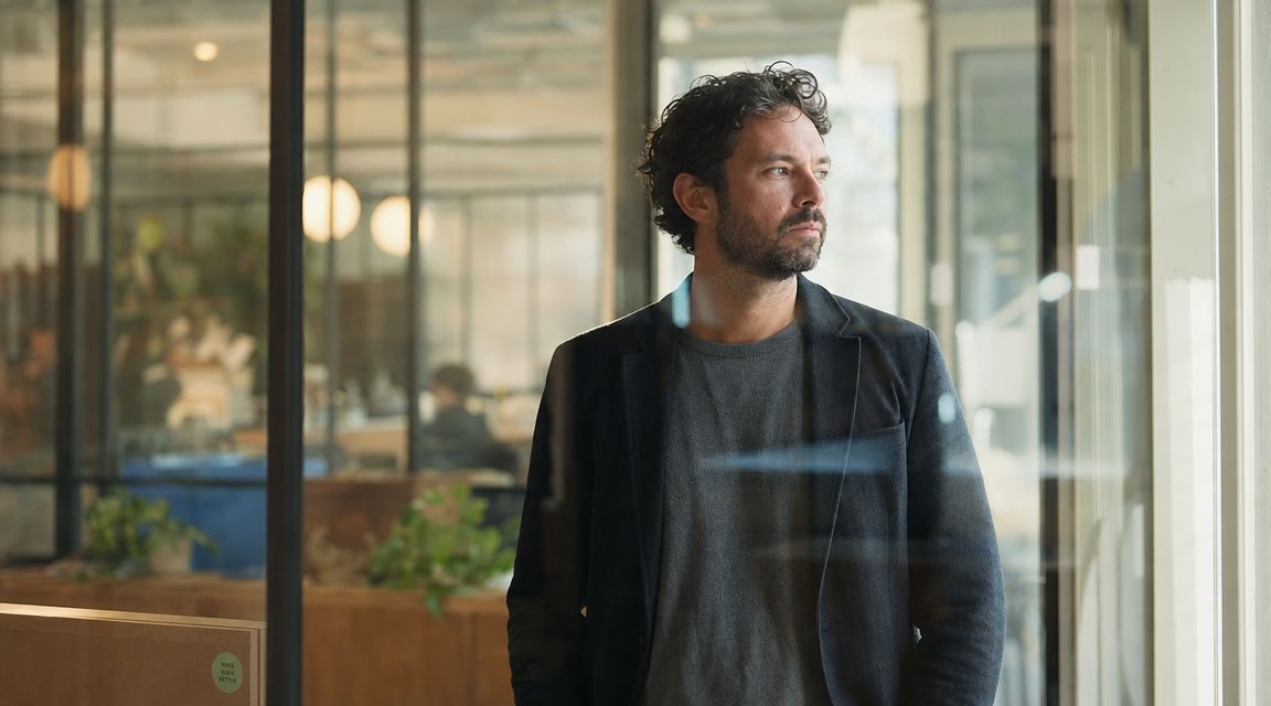 A thoughtful professional man in a business blazer looking out a window in a modern office space.