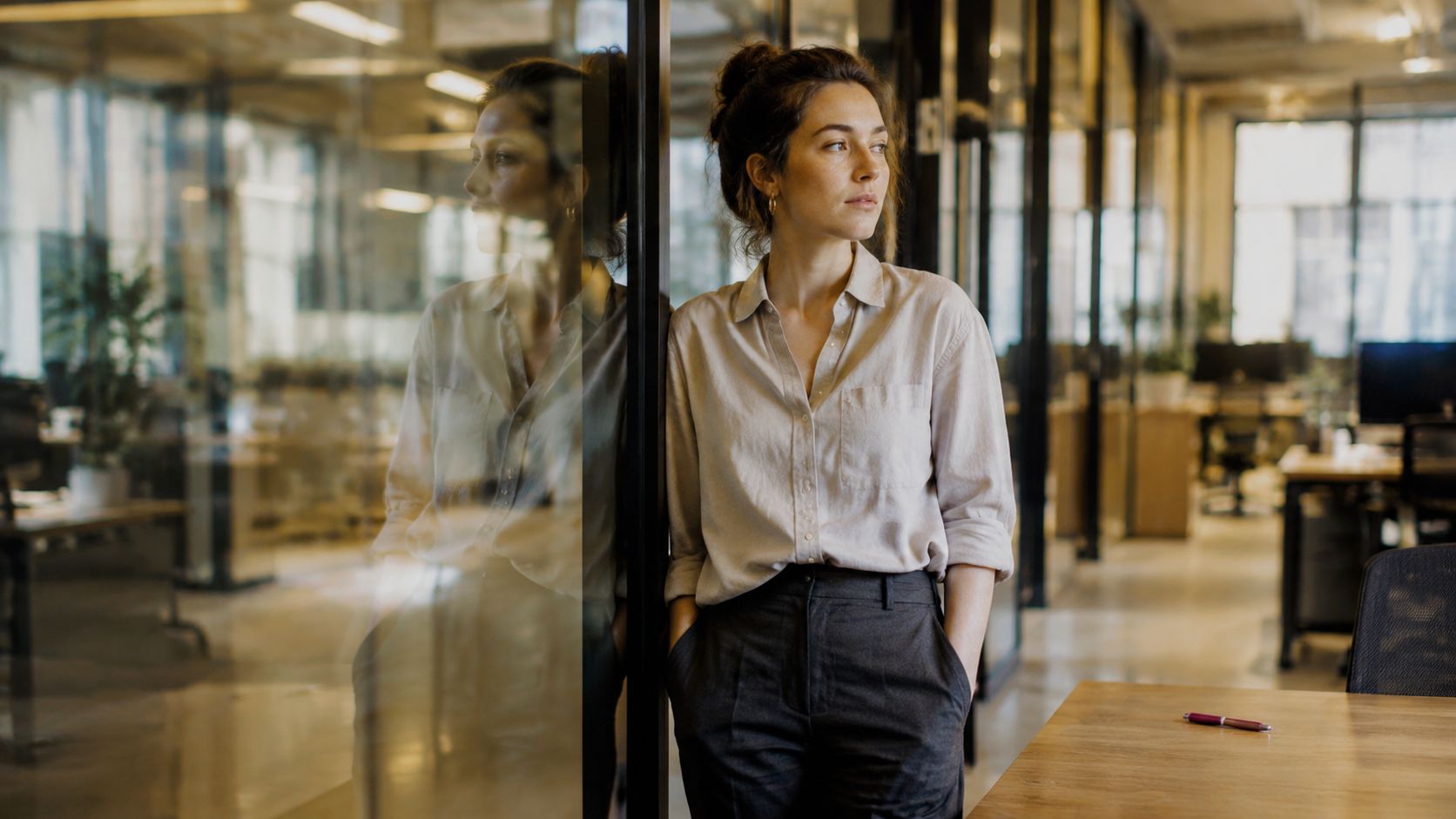 A professional woman in an office setting leaning against a glass partition with a thoughtful expression.