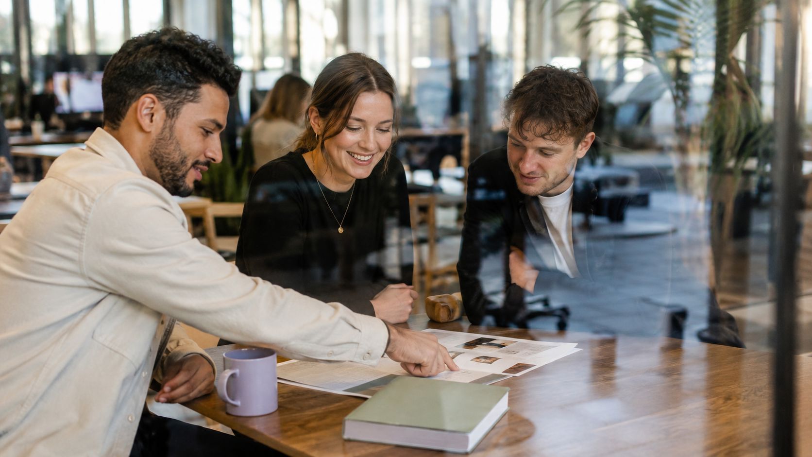 Three colleagues collaborate in a modern office, pointing at documents on a wooden desk together.