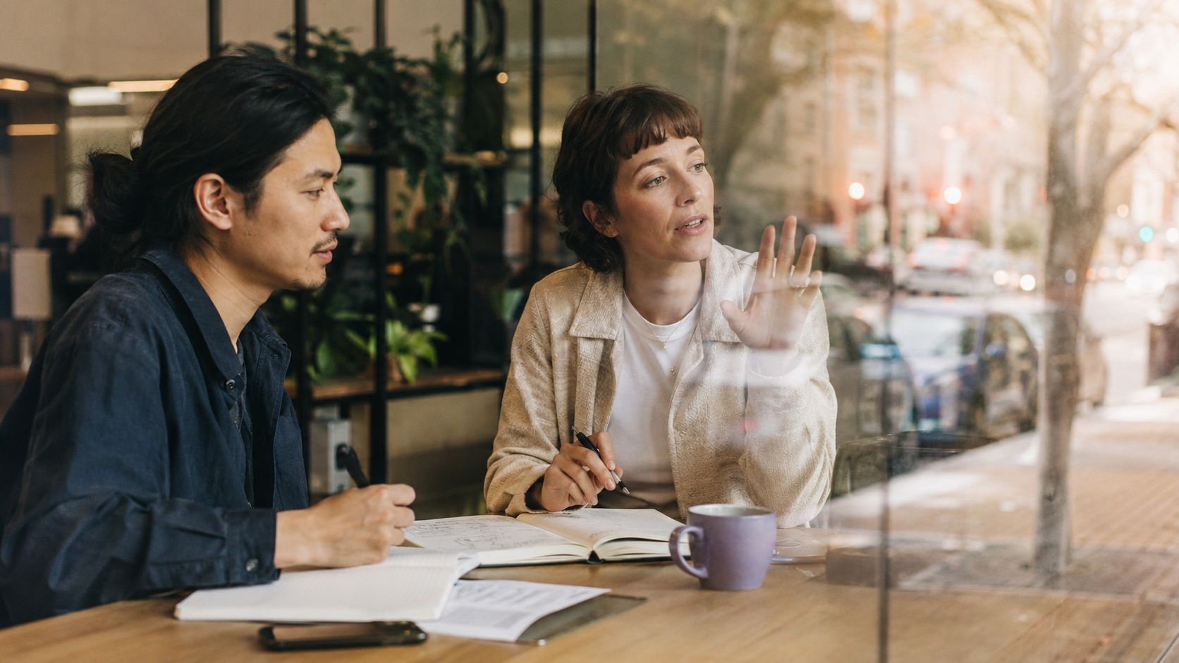 A professional man and woman collaborating on tasks with notebooks and a coffee cup in a cafe.