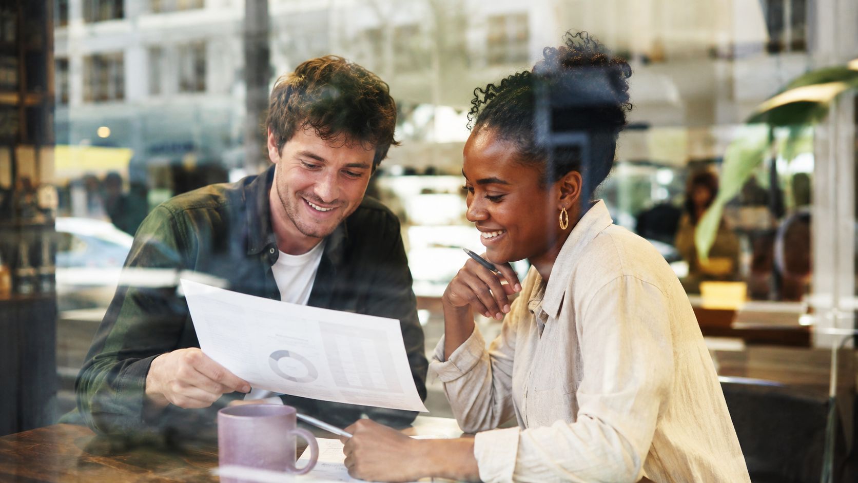 A diverse man and woman sitting at a cafe table reviewing business documents and financial charts together.