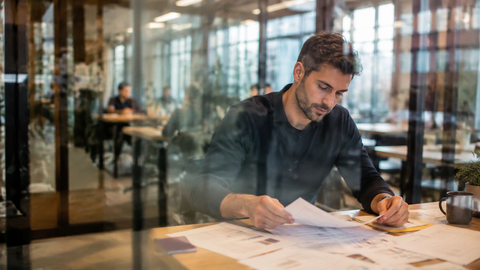 A professional man reviewing financial documents and business reports in a modern glass-walled office workspace.