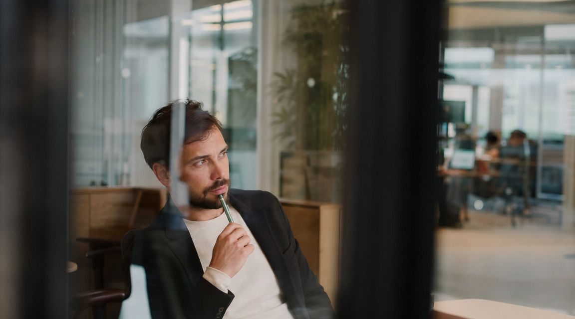 A professional man with a beard looking thoughtful while holding a pen in a modern office space.