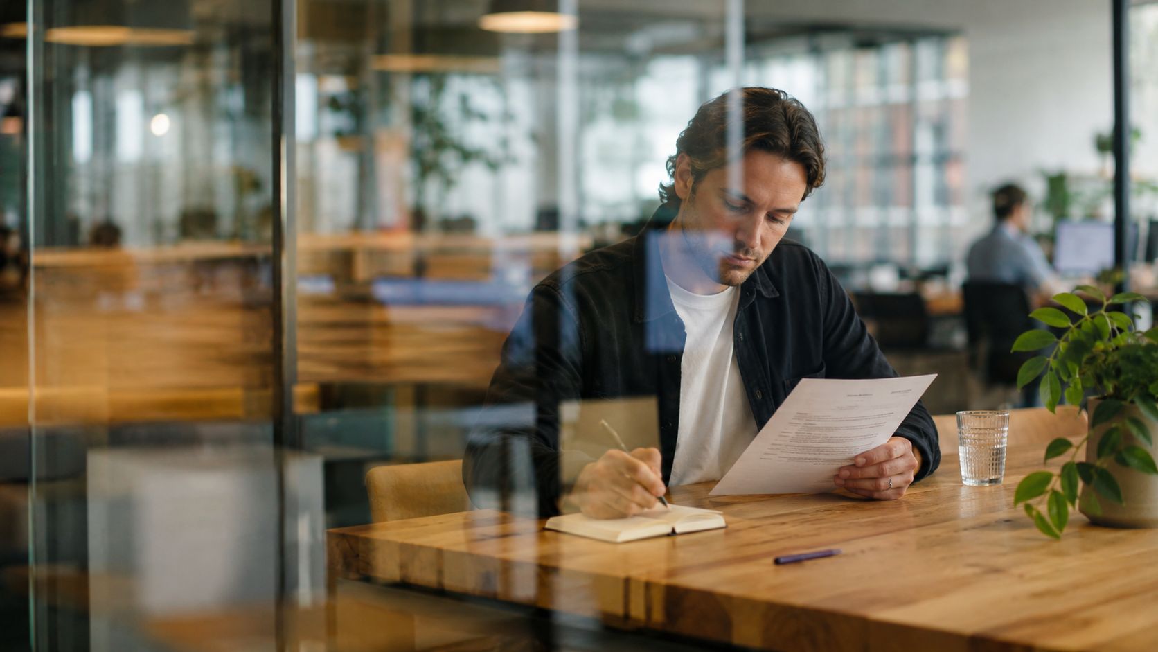 A professional man sitting at a wooden table in an office, reviewing a document and writing in a notebook.