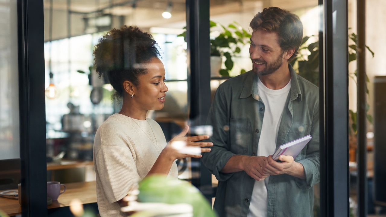 A professional woman and man discussing work while standing inside a bright, modern office space.