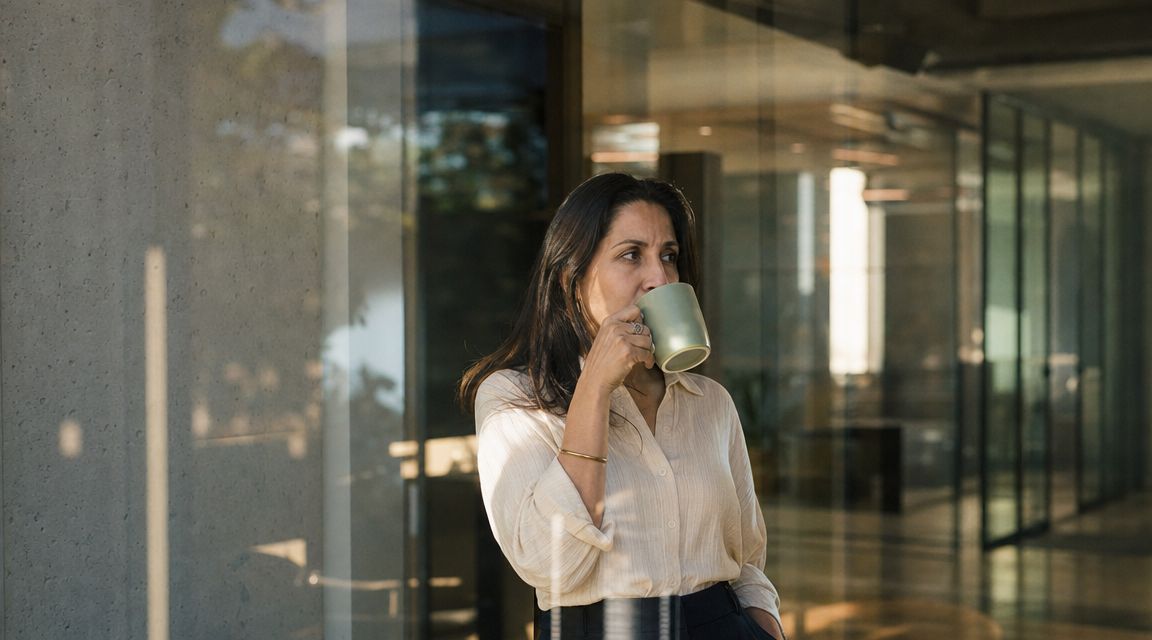 A professional woman drinking from a mug in a modern office with glass partitions and concrete walls.