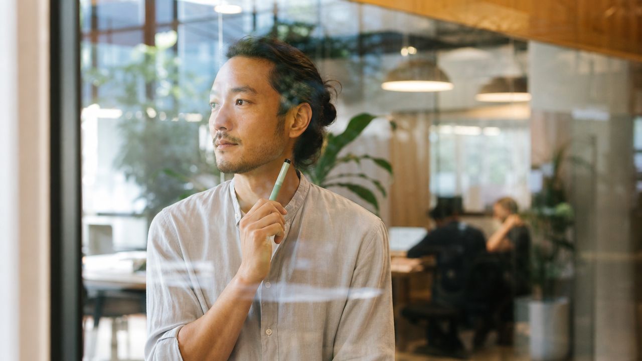 A thoughtful business professional holding a pen while looking through a glass partition in a modern office.