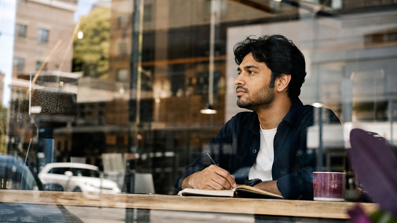 A focused man writing in a notebook while sitting by a window in a cafe setting.