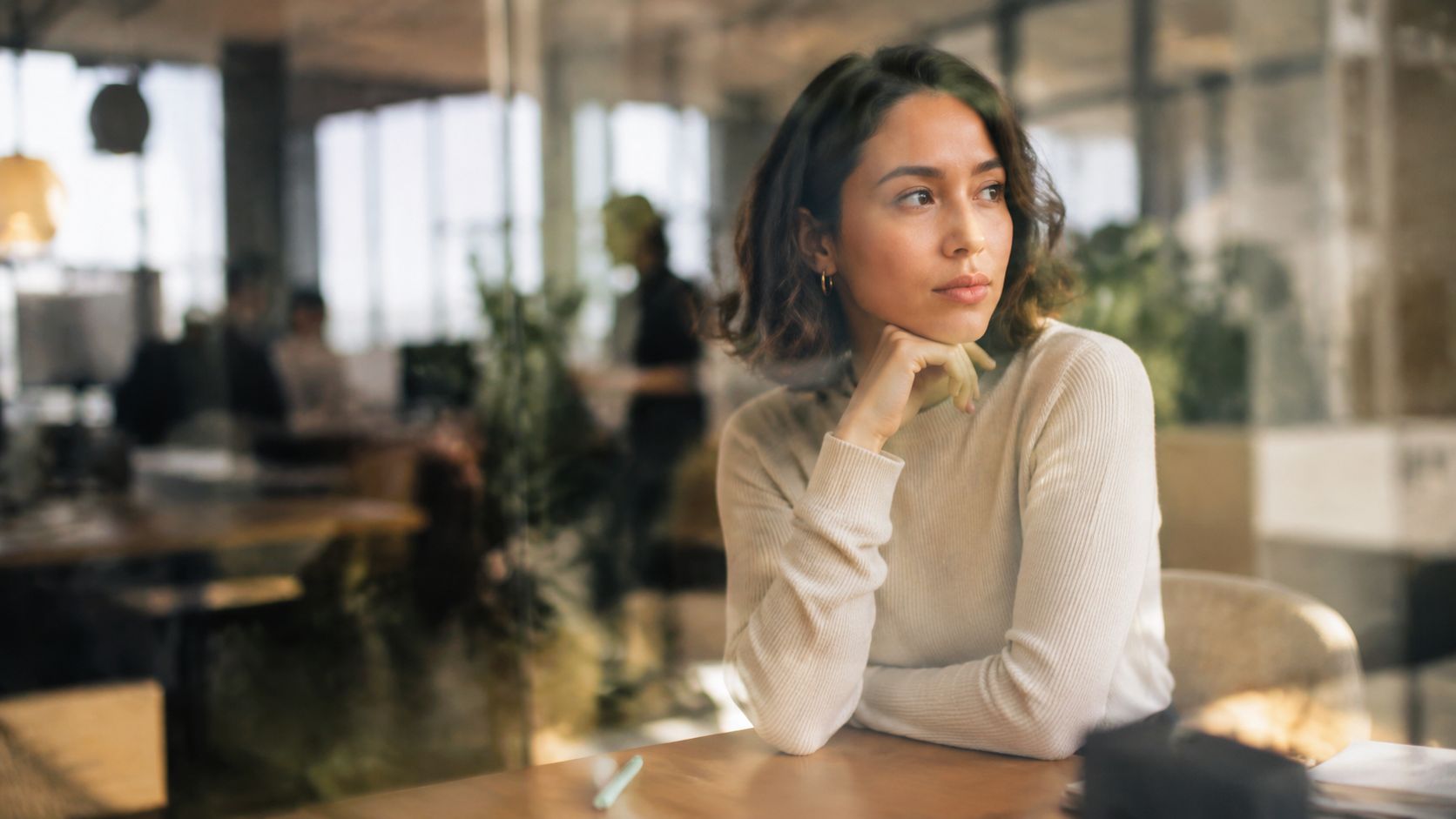 A professional woman in an office setting, sitting at a desk and thoughtfully looking to the side.