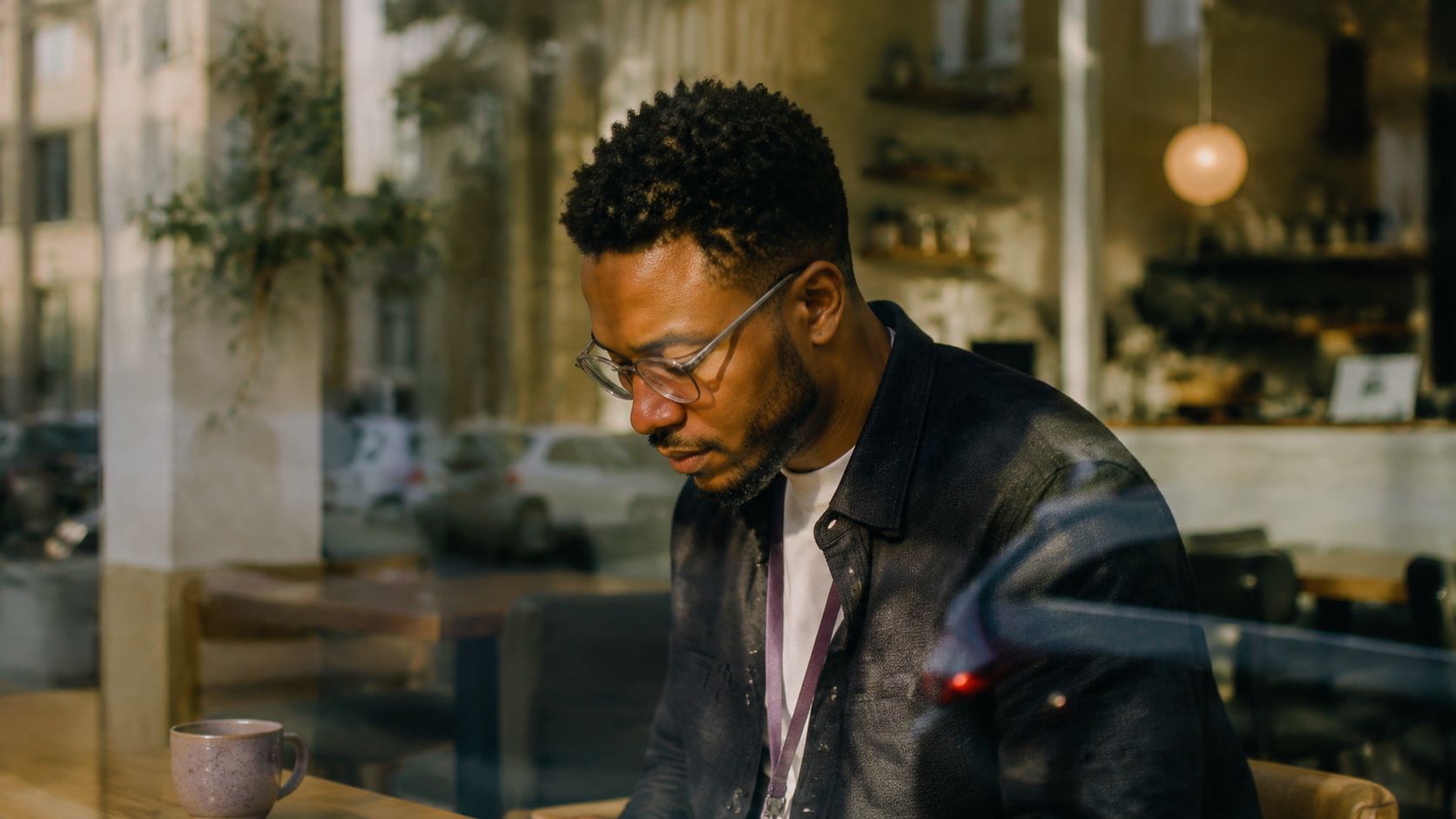 A young man with glasses working intently on his laptop while sitting in a cafe.