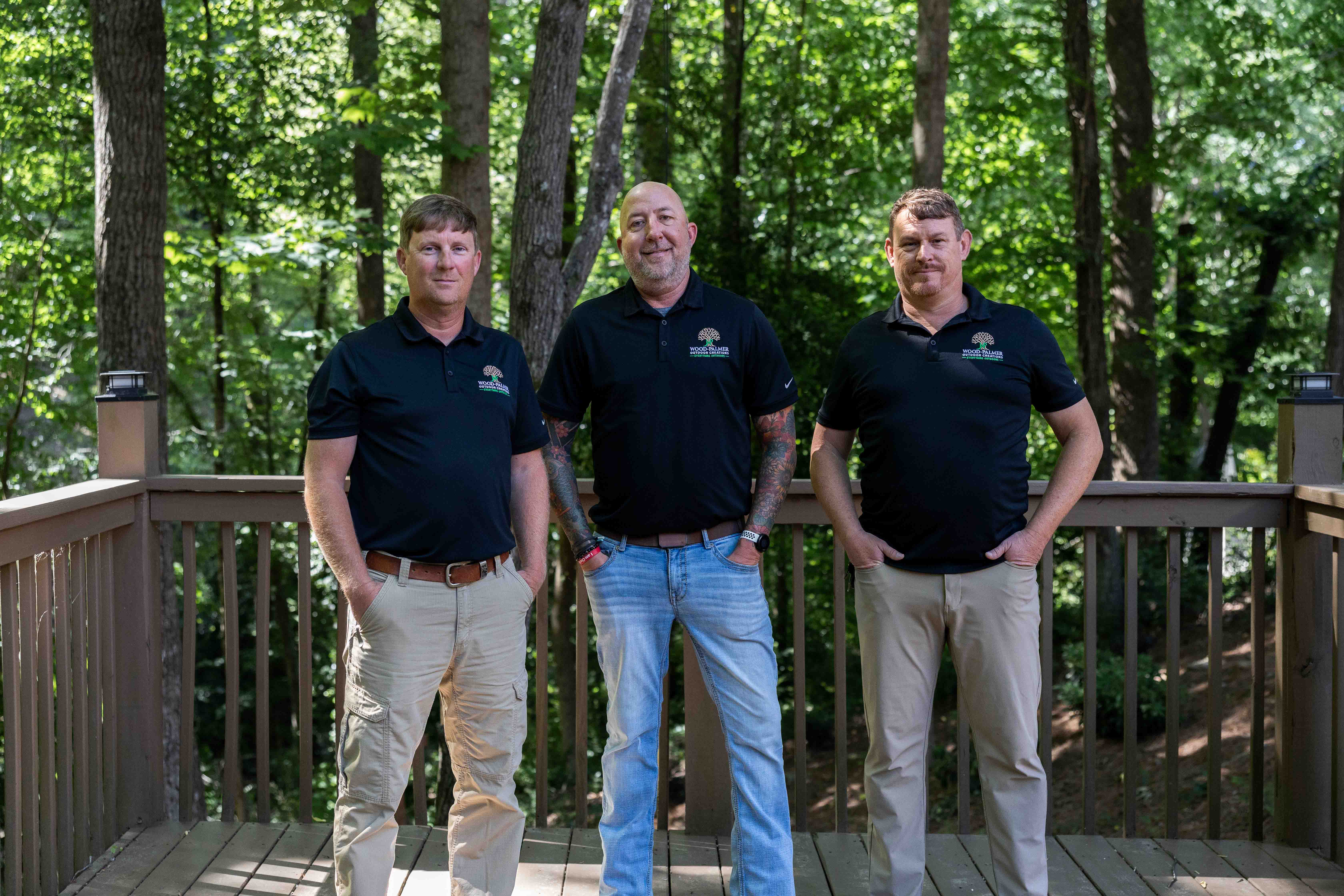 Three men wearing matching black polo shirts stand on a wooden deck with green trees behind them.
