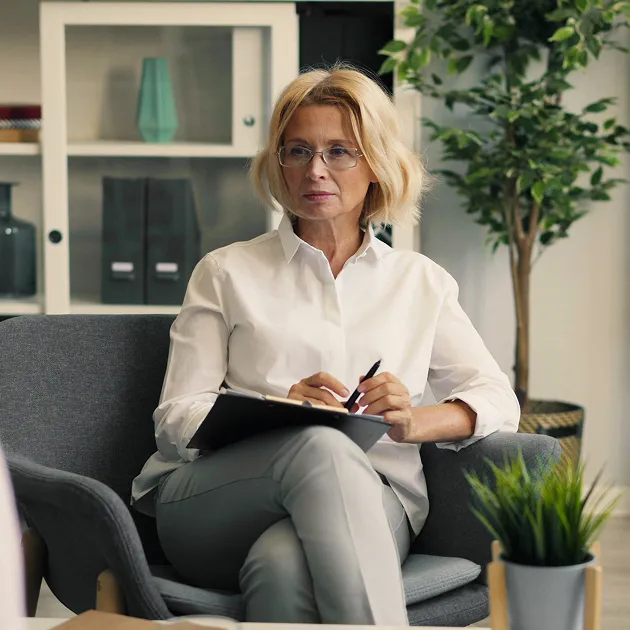 Woman with glasses holding a clipboard and pen, seated in a chair in a modern office setting.