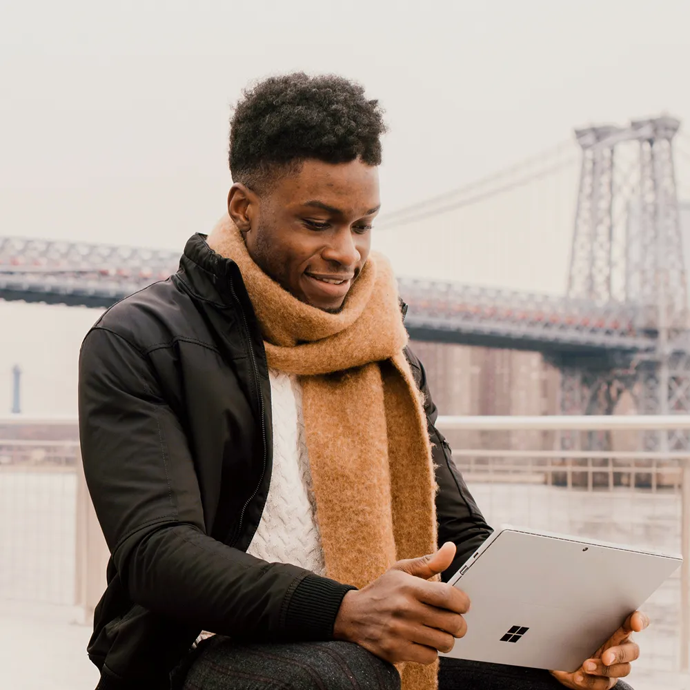 Man wearing a scarf and jacket using a tablet outdoors with a steel bridge in the background.