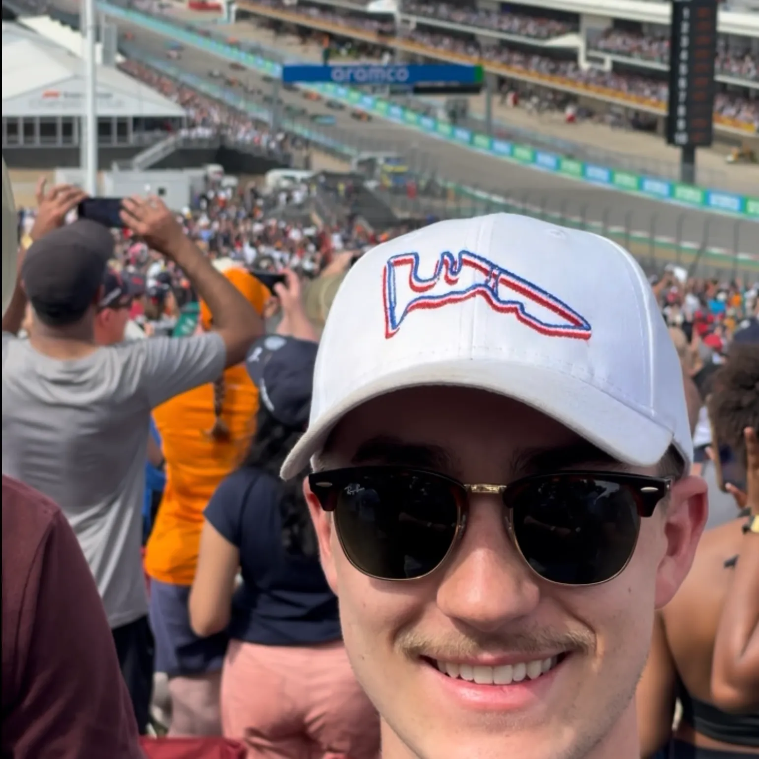 Young man wearing sunglasses and white cap at a crowded race track event with people taking photos.