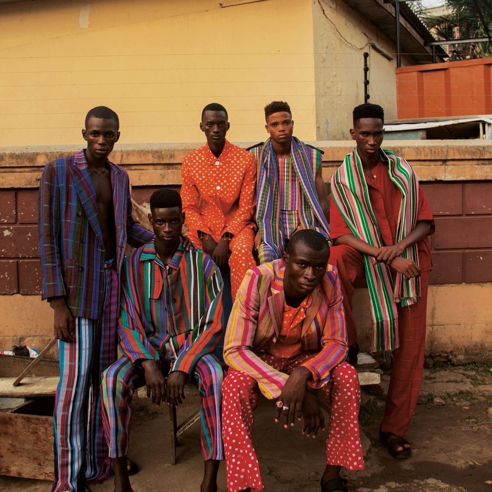 Six men dressed in vibrant, colorful patterned suits and striped scarves posing against a brick wall outdoors.