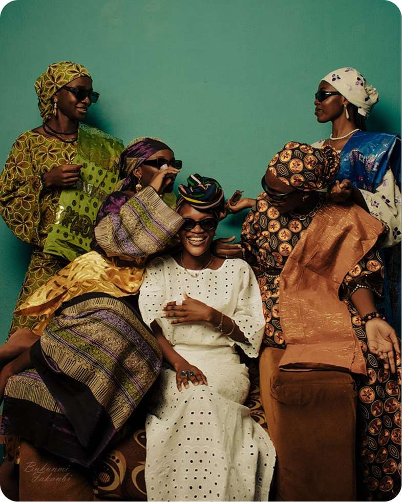 Group of five women wearing colorful traditional African attire and sunglasses, smiling and interacting against a teal background.