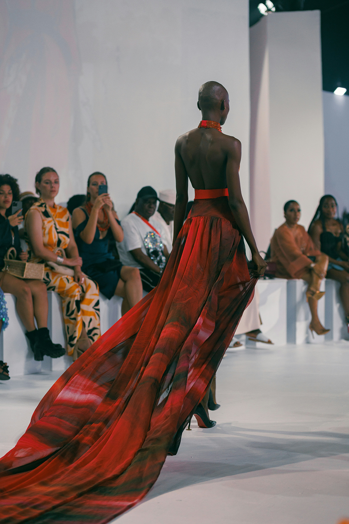 Model wearing a long flowing red dress walking on a runway during a fashion show with audience seated on both sides.