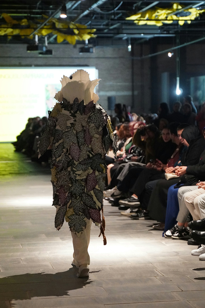 Model walking down runway wearing an outfit covered in large, textured leaf shapes, with an audience seated on both sides in a dimly lit industrial space.