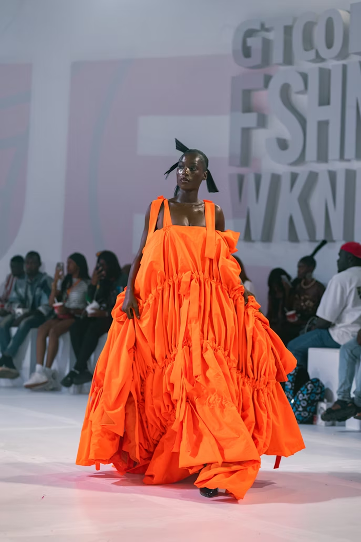 Model walking runway wearing voluminous bright orange dress with ruffled layers and wide straps.