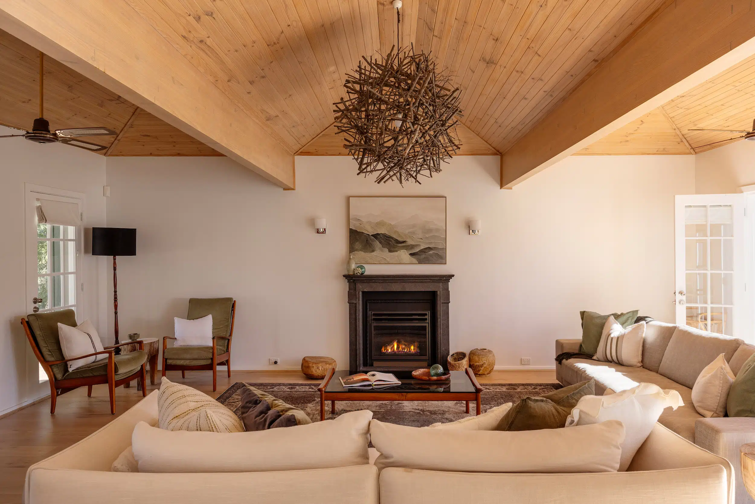 Cozy living room with beige sofas, green armchairs, lit fireplace, wooden ceiling, and twig chandelier.