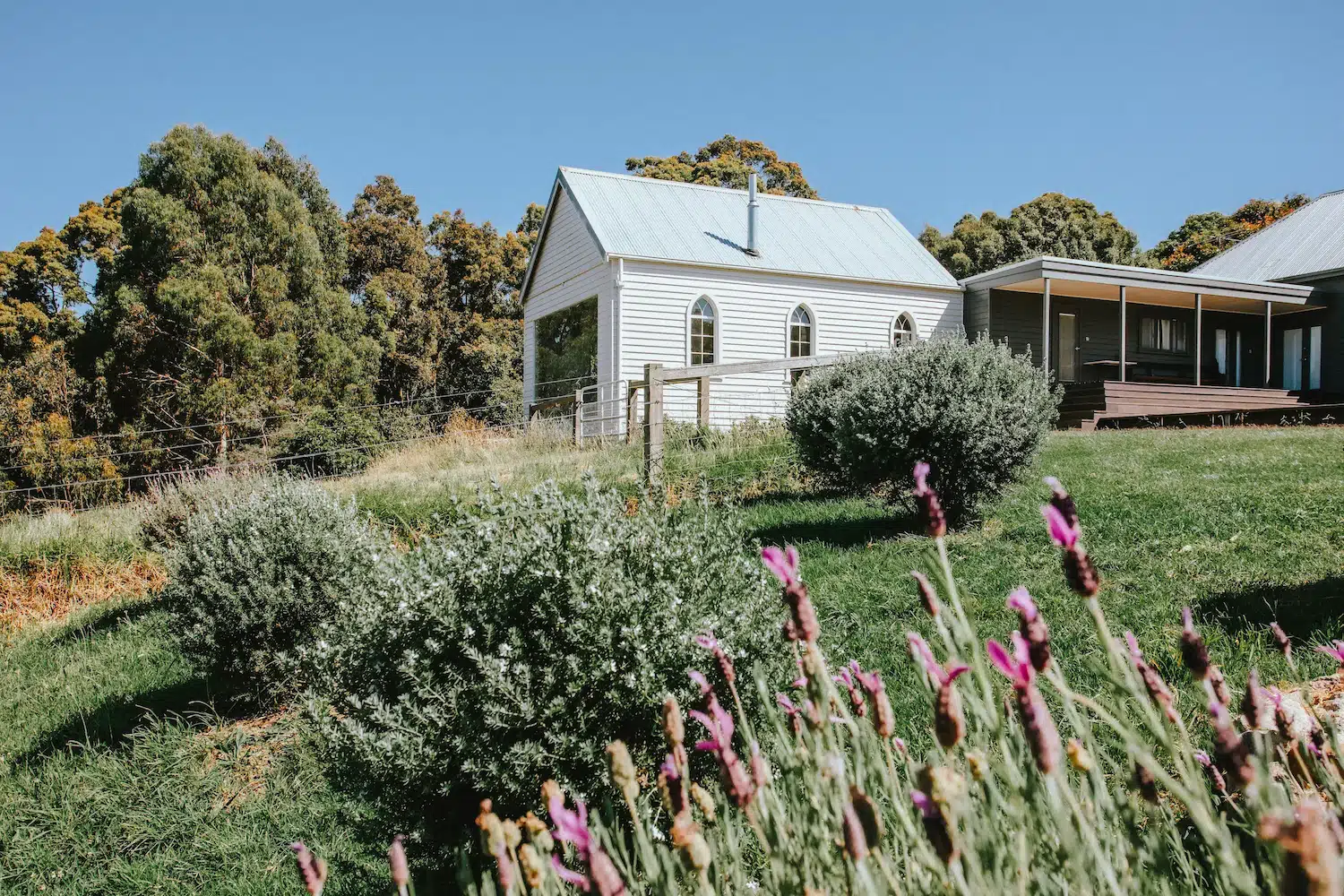 White cottage with arched windows on a grassy hill surrounded by bushes and purple flowers.