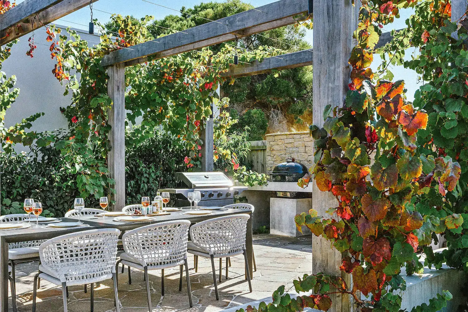 Outdoor dining table with white chairs under vine-covered wooden pergola and a grill in background.