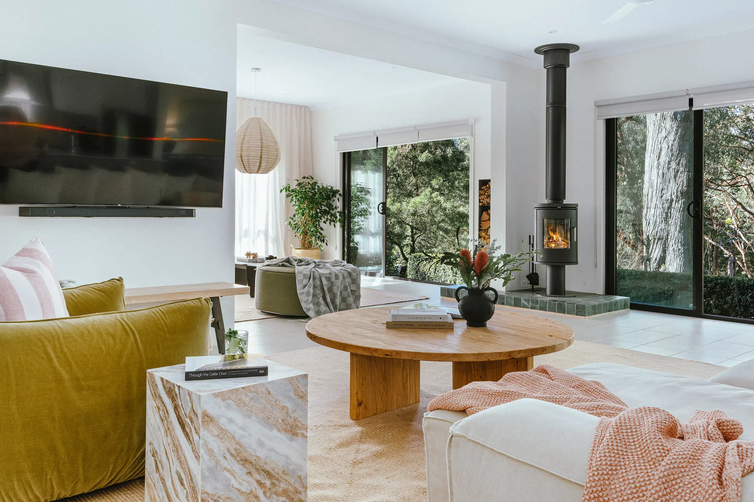 Modern living room with green sofa, round wooden coffee table, fireplace, and large windows showing trees.