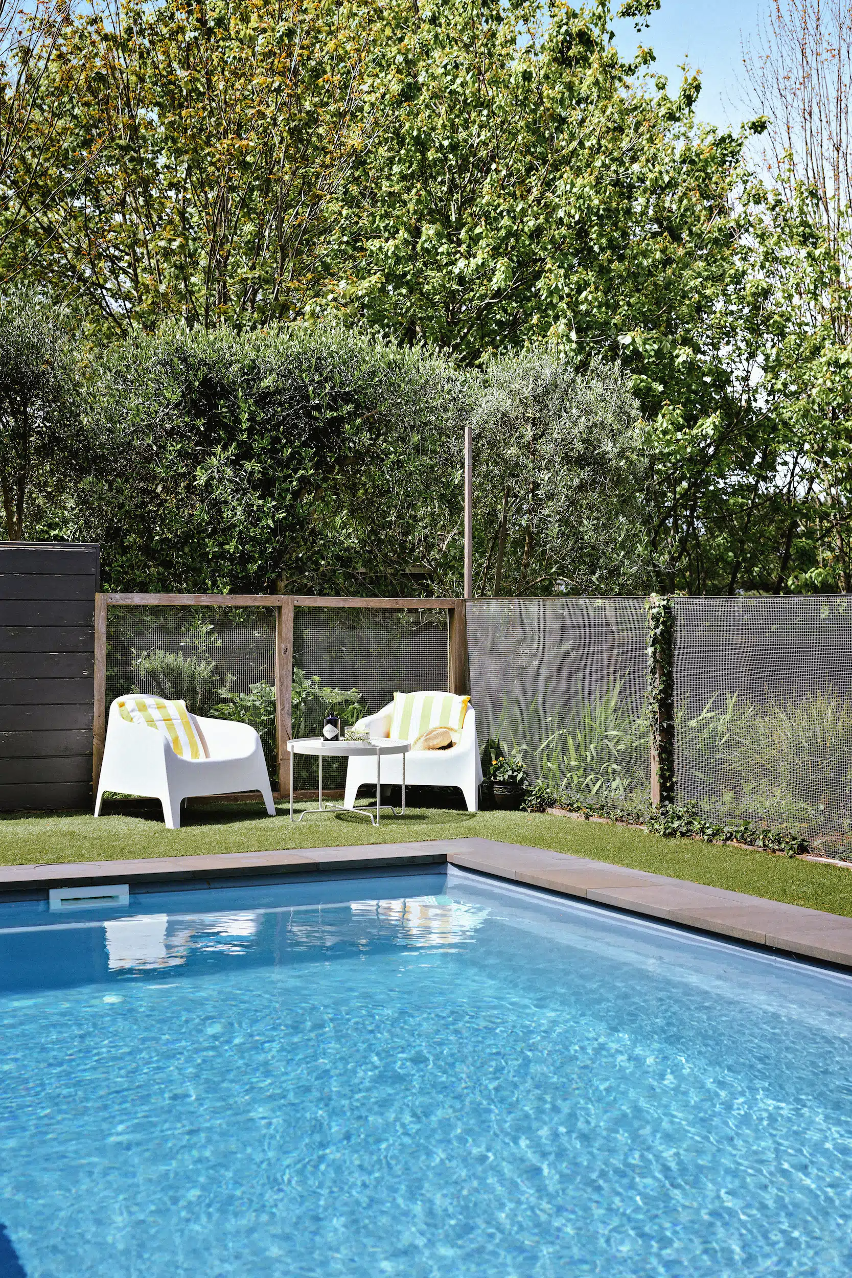 Blue outdoor pool with two white chairs and a table on grass beside a mesh fence and green bushes.