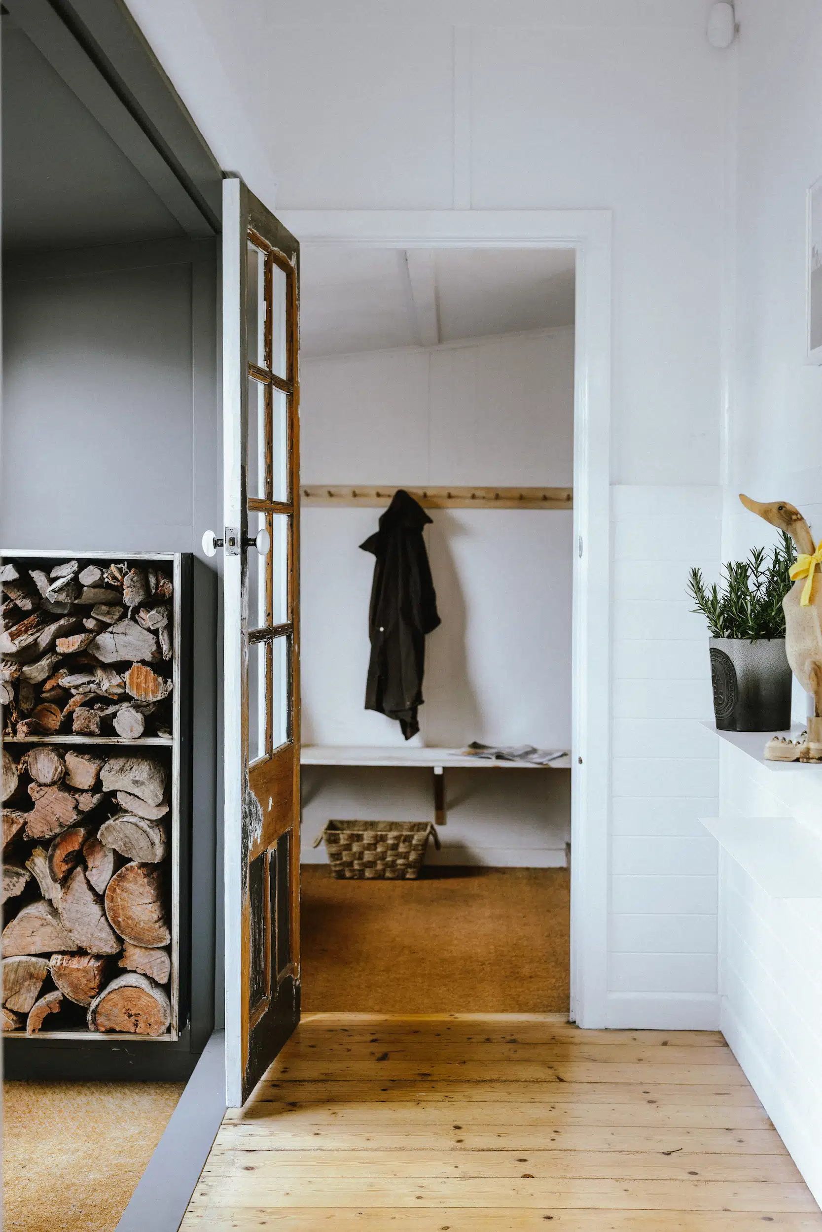 Open door shows a coat hanging on a rack, firewood stacked inside, and a plant with a duck figurine.