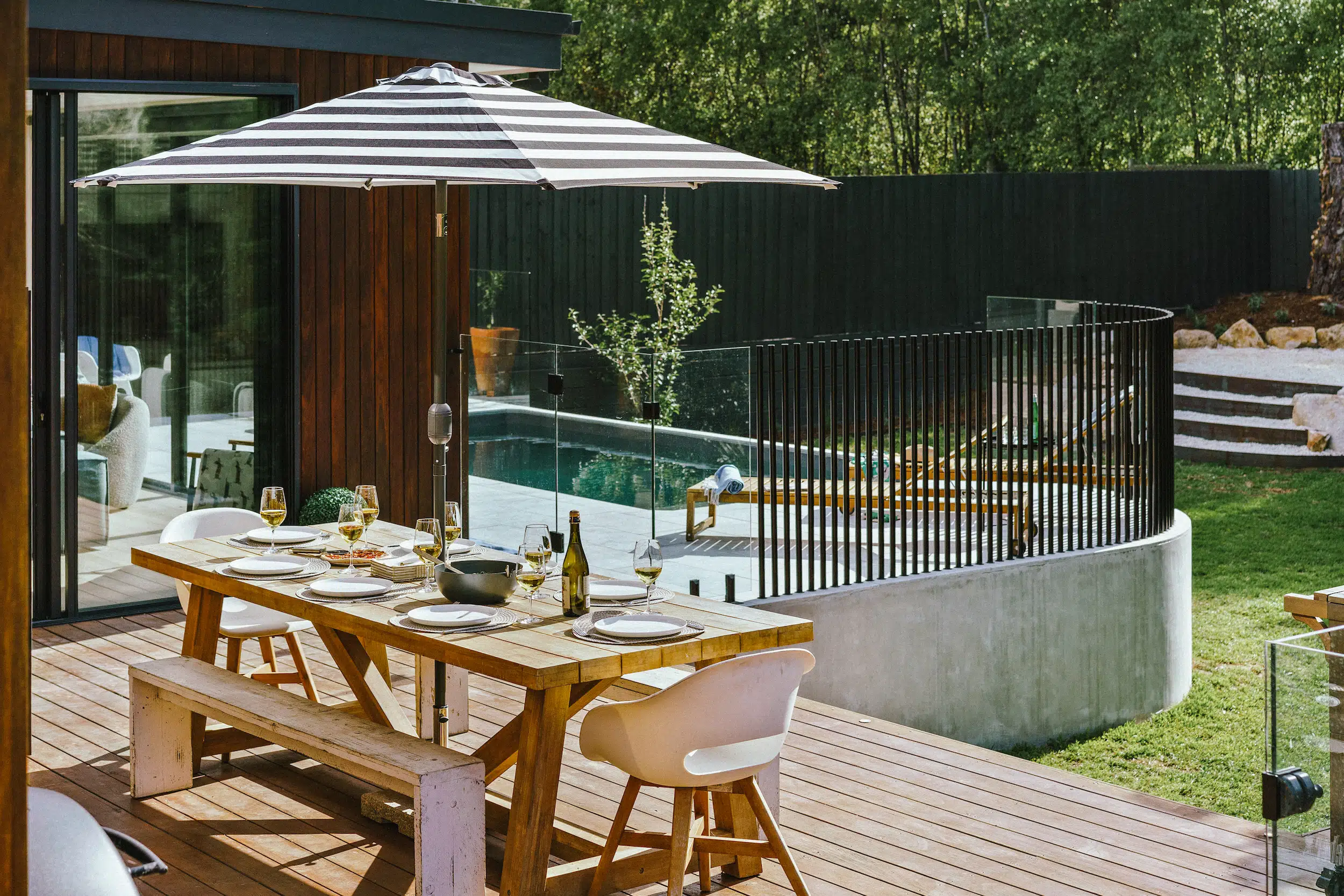 Outdoor wooden dining table with white plates and wine glasses under a striped umbrella on a deck.